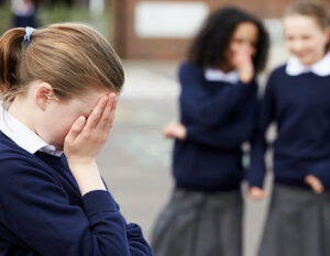 An image of a girl with her face in her hands and two girls whispering behind her in the playground