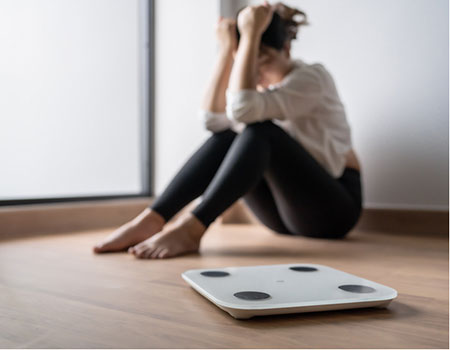 An image of a girl sat on the floor looking frustrated next to bathroom scales